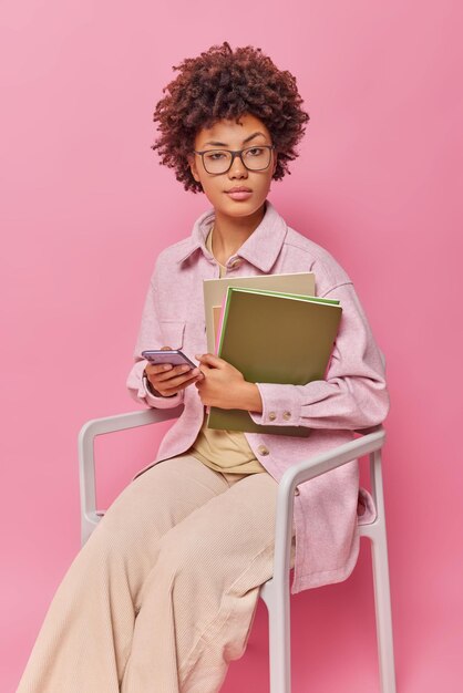 Clever female student holds mobile phone and notepads wears transparent glasses casual clothes studies distantly prepares for exam sits on chair isolated over pink wall.