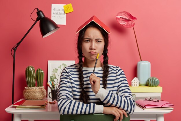 Clever korean schoolgirl has serious sullen look, folds lips, holds pencil, notepad on head