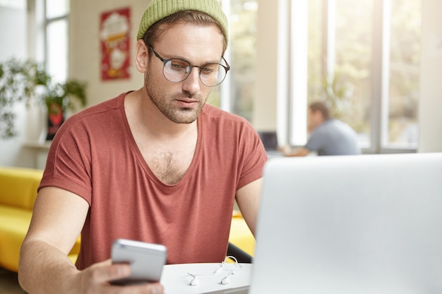 Clever male office worker sits at coffee shop, wears spectacles and fashionable clothes