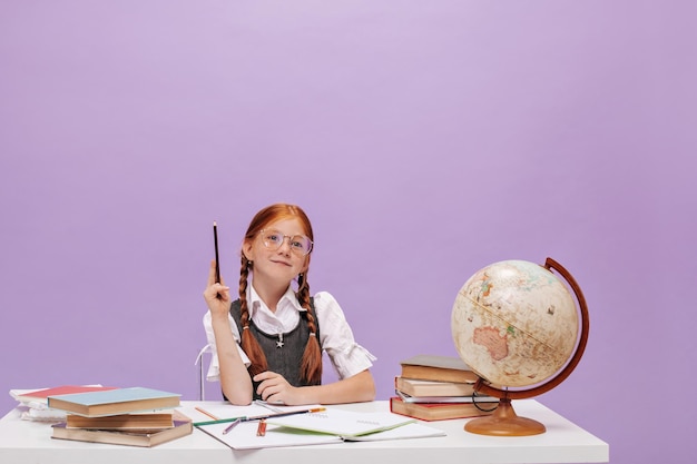 Clever red haired small girl with modern hairstyle in clear eyeglasses and white blouse wants to answer and raises her hand