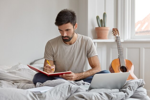 Clever student copies information from webpage into notepad, does homework, makes notes, sits at bed near opened laptop computer, has serious expression. Busy male copywriter records good ideas.