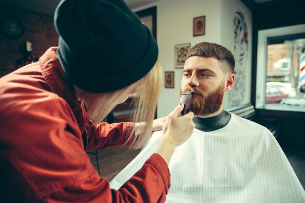 Client during beard shaving in barbershop. Female barber at salon. Gender equality