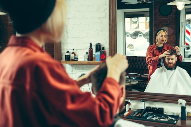 Client during beard shaving in barbershop. Female barber at salon. Gender equality