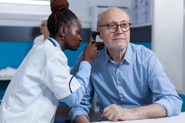 Clinic otology specialist consulting senior patient using otoscope to check ear infection. Hospital otologist examining sick retired man internal ear condition while in doctor cabinet.