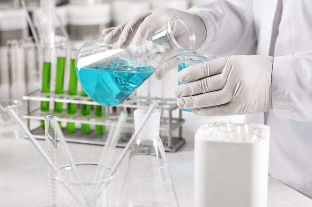 Clinical worker dressed in white gown and gloves holding glass beakers with blue liquid