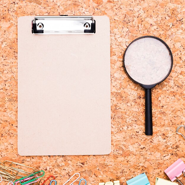 Clipboard with magnifier and multicolored paper clips on cork background