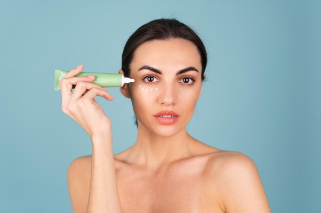 Close beauty portrait of a topless woman with perfect skin and natural make-up, holding a cream for moisturizing and elasticity of the skin under the eyes