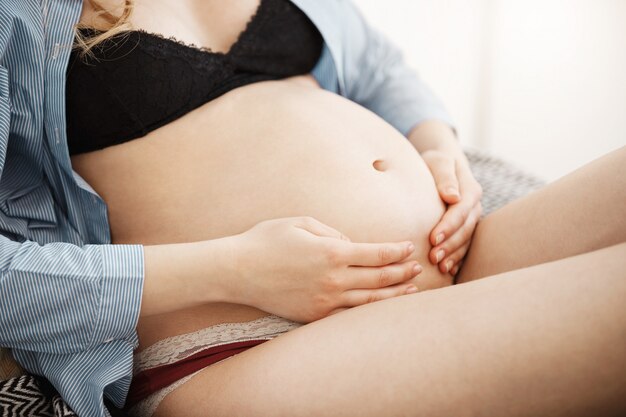 행복하게 손으로 그녀의 임신 배꼽을 잡고 목욕 가운 아래 검은 란제리에 젊은 어머니의 자른보기를 닫습니다. 여자의 순수한 사랑의 달콤한 장면. 출산 개념