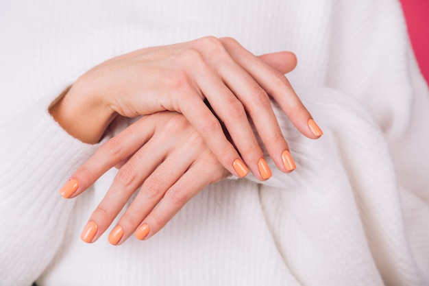 Close frame of woman's hands with orange manicure on white sweater and pink wall
