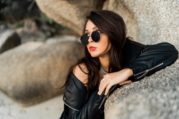 Close op portrait of confident woman with red lips in trendy black leather jacket and sensual black dress posing on rocky background