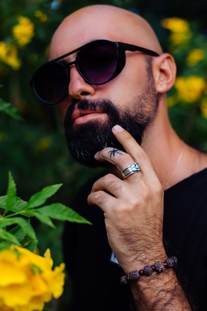 Close shot of brutal tanned bearded man in sunglasses having palm tree tattoo on finger stands surrounded by yellow flowers in park