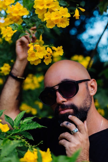 Close shot of brutal tanned bearded man in sunglasses having palm tree tattoo on finger stands surrounded by yellow flowers in park