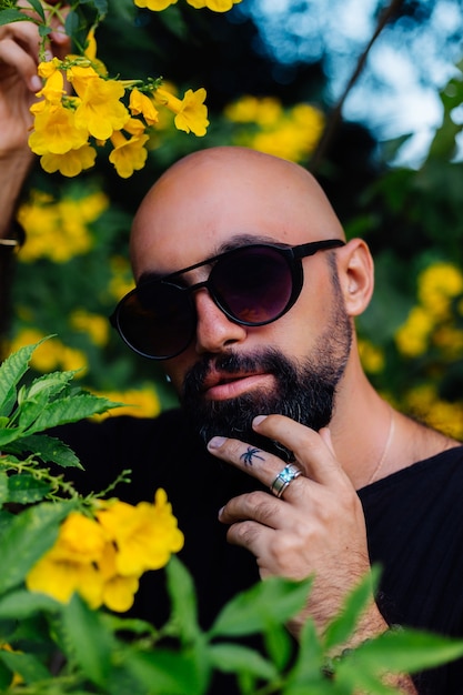 Close shot of brutal tanned bearded man in sunglasses having palm tree tattoo on finger stands surrounded by yellow flowers in park