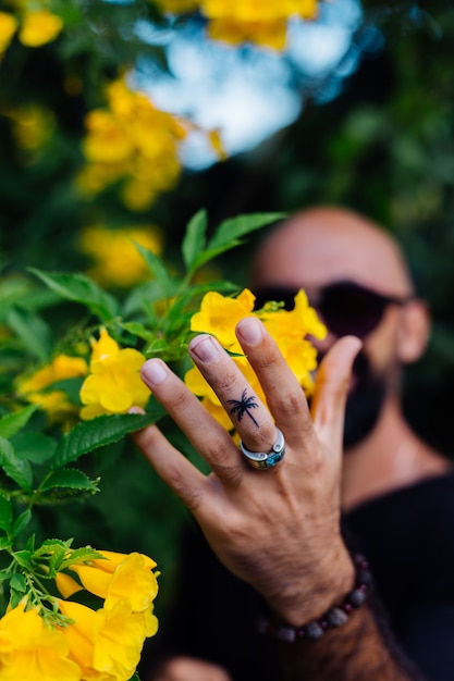 Close shot of brutal tanned bearded man in sunglasses having palm tree tattoo on finger stands surrounded by yellow flowers in park