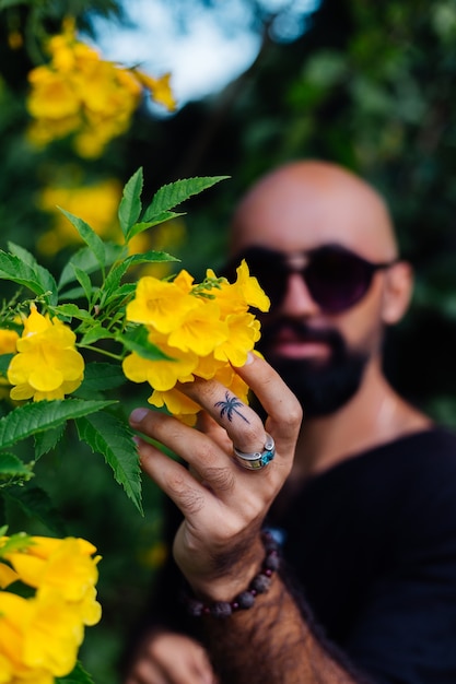 Close shot of brutal tanned bearded man in sunglasses having palm tree tattoo on finger stands surrounded by yellow flowers in park