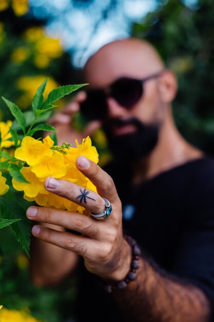 Close shot of brutal tanned bearded man in sunglasses having palm tree tattoo on finger stands surrounded by yellow flowers in park