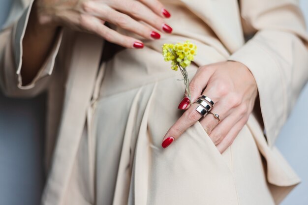 Close shot of woman hands red manicure two rings wearing beige suit. Yellow cute dried flower in pocket.