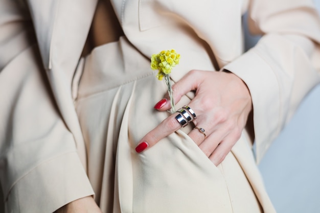 Close shot of woman hands red manicure two rings wearing beige suit. Yellow cute dried flower in pocket.