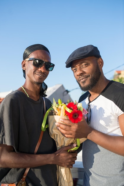 Primo piano delle coppie gay africane che scattano foto con i fiori. due uomini sorridenti felici in piedi con calma vicino tenendo il mazzo di bellissimi fiori in posa guardando la fotocamera. vita delle coppie lgbt, concetto di relazioni