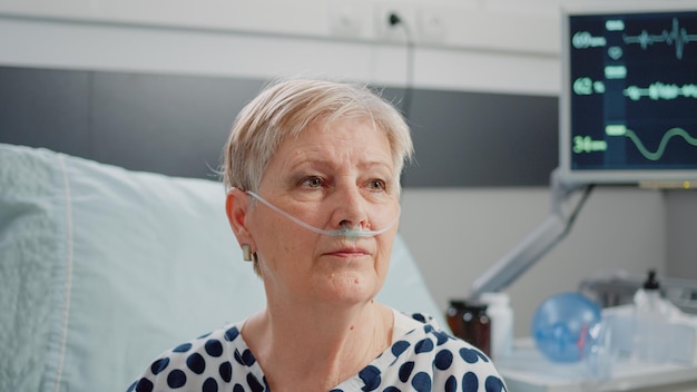 Close up of aged woman sitting in bed with heart rate monitor at clinic. Portrait of patient with illness using nasal oxygen tube for respiratory healthcare and recovery. Sick pensioner