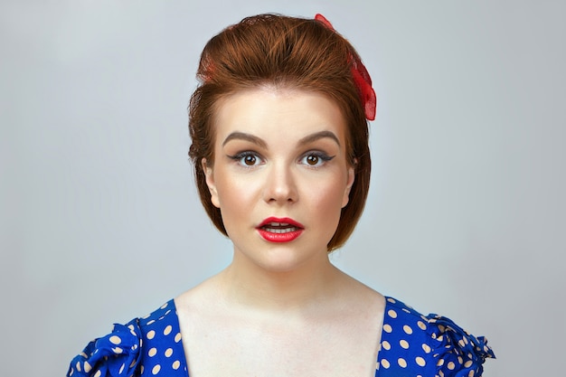 Free photo close up of amazed pinup girl wearing bright make up, vintage dress and retro hairstyle staring at camera in shock with jaw dropped