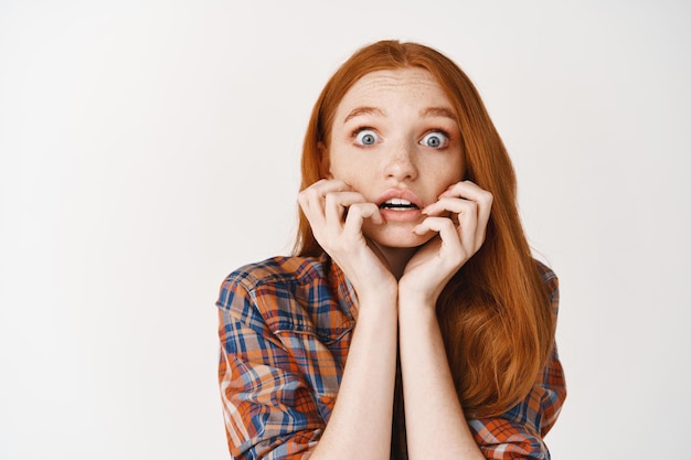Close-up of amazed redhead girl listening with interesting, biting fingernails tempted and amused, looking at front, standing over white wall