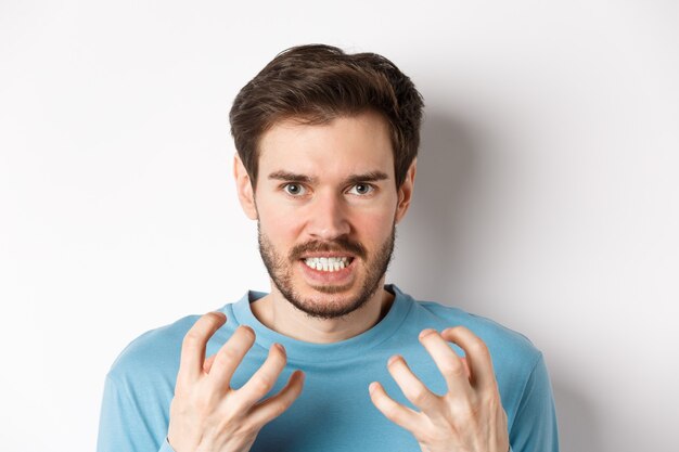 Close up of angry young man with beard, shaking hands mad, squeeze teeth and frowning furious, standing outraged over white background