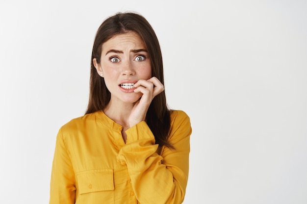 Close-up of anxious young woman biting fingernails, staring at camera insecure and nervous, standing unconfident on white wall.