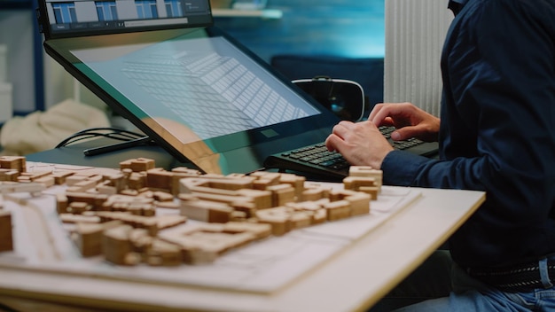 Close up of architect hands using touch screen computer and maquette on table. Man engineer working with technology to analyze building model for construction layout and development