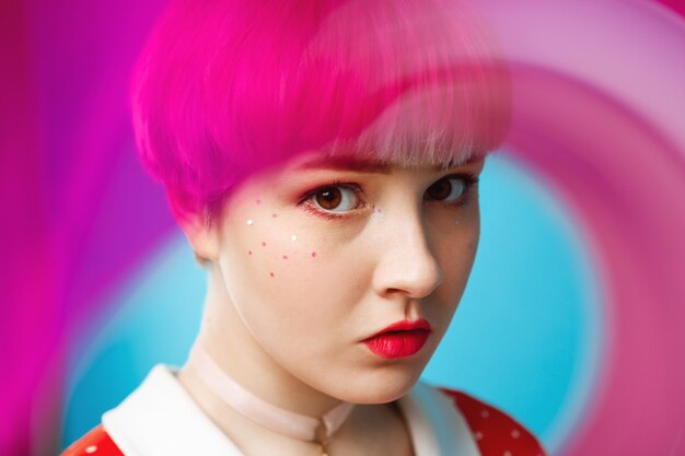 Close up artistic conceptual portrait of beautiful dollish girl with short light violet hair wearing red dress over blue wall Blurry forefront.