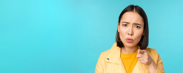 Close up of asian girl accusing and pointing finger at camera looking offended or displeased scolding you standing over blue background