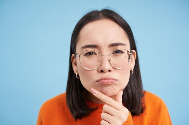 Close up of asian woman in glasses thinking staring at camera with thoughtful face expression standi