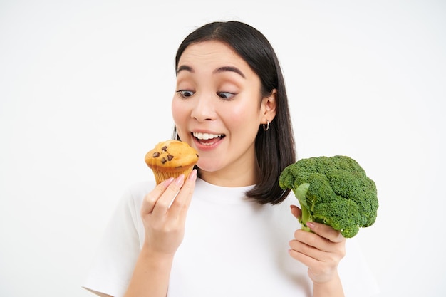 ブロッコリーを保持しているが、おいしいカップケーキを食べるアジアの女性のクローズ アップ ホワイト スタジオの背景