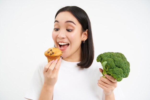 Крупный план азиатской женщины, держащей брокколи, но поедающей вкусный кекс на белом студийном фоне