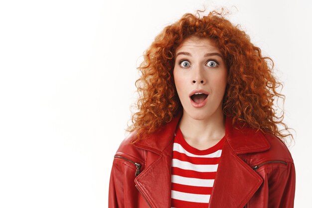 Close-up astonished surprised excited redhead curly-haired girl blue eyes wearing stylish modern red jacket drop jaw wondered widen eyes amazed reacting excitement awesome news white wall