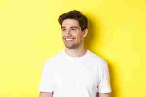 Free photo close-up of attractive bearded man in white t-shirt smiling, looking left at copy space, standing against yellow background.