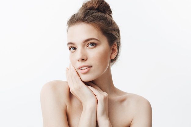 Close up of attractive dark-haired young woman with bun hairstyle and naked shoulders holding hands near face,  with relaxed and calm face expression.