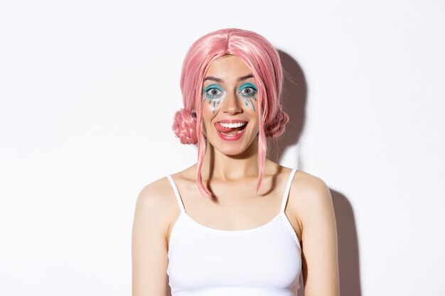 Close-up of attractive happy girl showing tongue and smiling, wearing pink party wig and bright makeup, standing.