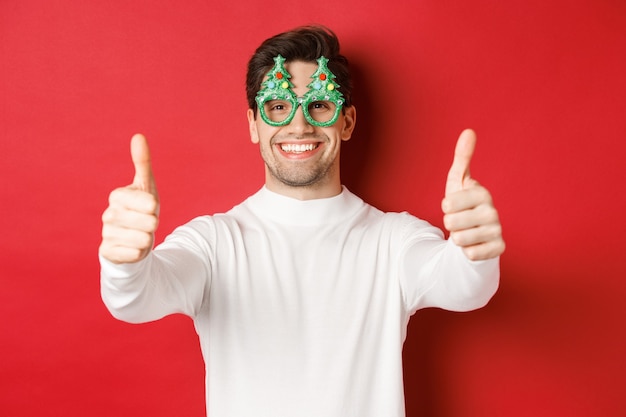 Primo piano dell'uomo felice attraente in occhiali da festa e maglione bianco, mostrando il pollice in su in segno di approvazione e sorridente, in piedi su sfondo rosso.