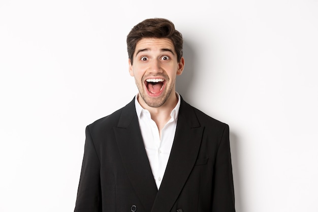 Close-up of attractive man in black suit, smiling amazed and looking at advertisement, standing over white background