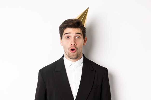 Close-up of attractive man in party hat and trendy suit, looking surprised, celebrating new year, standing against white background