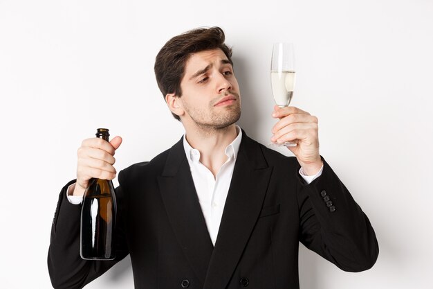 Close-up of attractive man in trendy suit, tasting champagne, looking at glass, standing against white background.