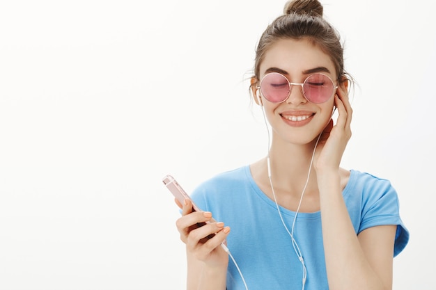 Close-up of attractive modern woman in sunglasses listening music, enjoying song in earphones, holding smartphone