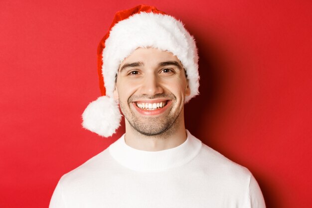 Close-up of attractive smiling man in white sweater and santa hat, looking happy, enjoying winter holidays, standing against red background