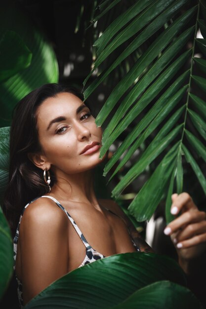 Close up of attractive young woman with perfect tan wearing bikini posing in tropical jungle palm leaves with sensual smile Female tourist relaxing on vacation