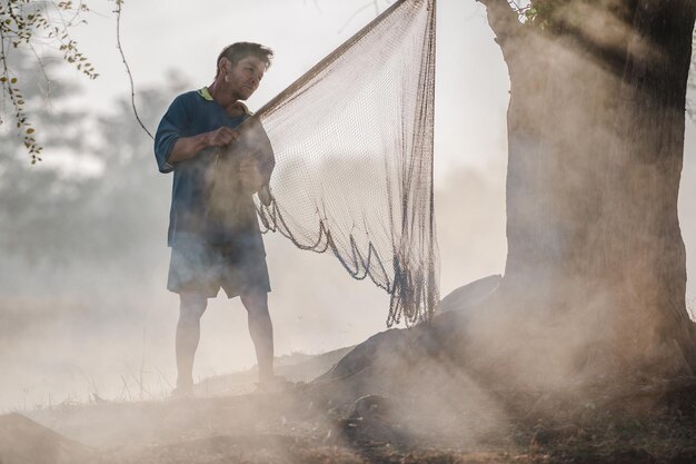 Крупным планом, сцена с подсветкой Азиатский рыбак проверяет или ремонтирует рыболовную сеть под деревом у реки рано утром с дымом и солнечным светом, пространство для копирования