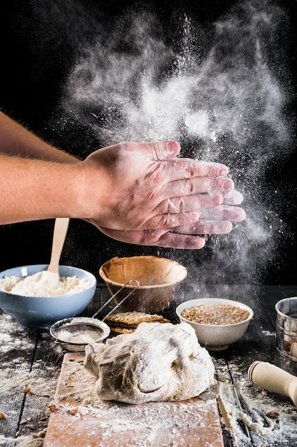 Primo piano della farina della spolverata della mano del panettiere sulla pasta con gli ingredienti sulla tavola