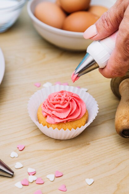 Close-up of bakers hand piping butter cream frosting on cupcake over the wooden desk