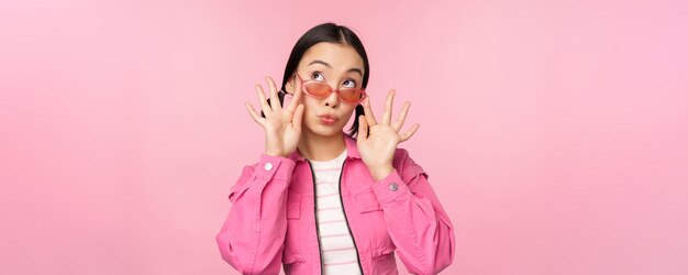 Close up of beautiful asian female model in stylish sunglasses posing against pink background in trendy outfit copy space