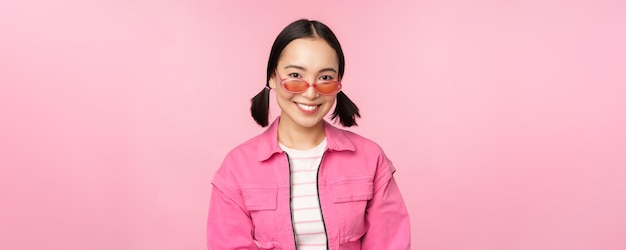 Крупный план красивой азиатской модели в стильных солнцезащитных очках, позирующей на розовом фоне в модном копировальном пространстве